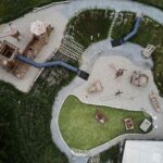 Luftaufnahme eines Spielplatzes mit Holzkonstruktionen, Rutschen, Klettergerüsten, Kieswegen und einem kleinen grünen Teich, der von Gras und Vegetation umgeben ist.