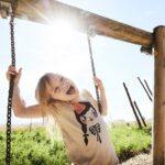 Ein kleines Kind, das sich an den Ketten einer Schaukel festhält, lächelt mit offenem Mund. Die Sonne scheint hell hinter ihnen auf einem Spielplatz im Freien.