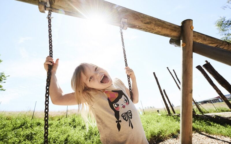 Ein kleines Kind, das sich an den Ketten einer Schaukel festhält, lächelt mit offenem Mund. Die Sonne scheint hell hinter ihnen auf einem Spielplatz im Freien.