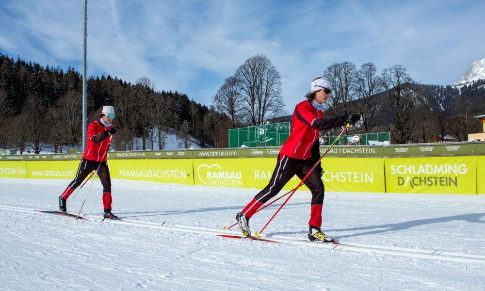 Zwei Personen in roter Sportkleidung beim Skilanglauf auf einer verschneiten Piste mit einem grünen und gelben Werbebanner im Hintergrund.