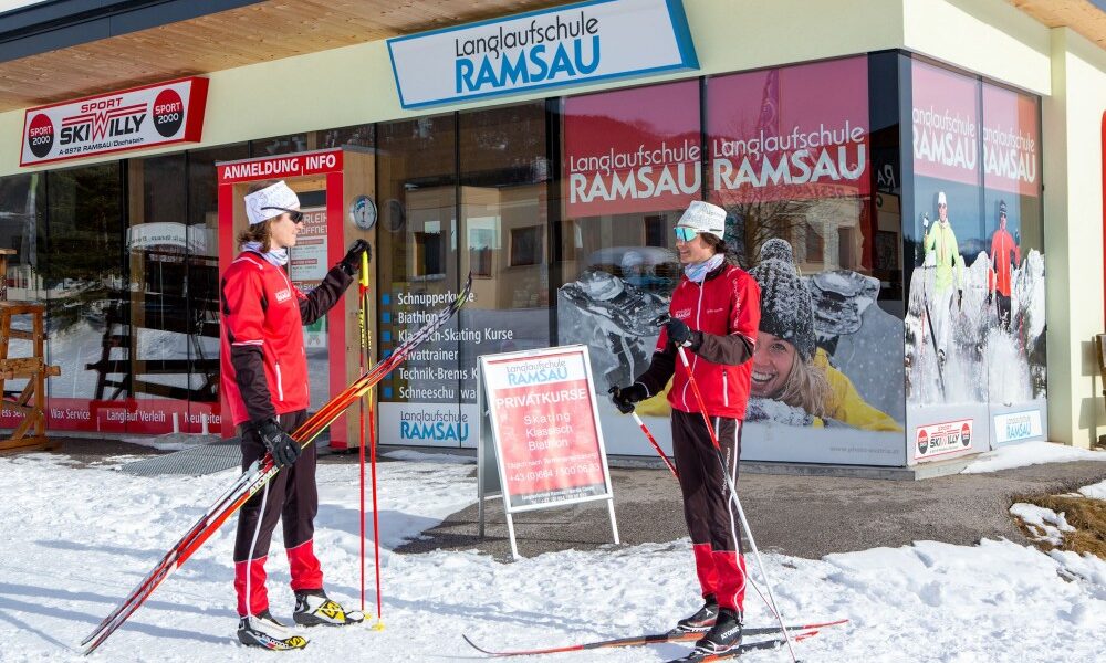 Zwei Personen in roter Skikleidung stehen mit Skiern vor dem Gebäude einer Langlaufschule, auf dem Boden liegt Schnee und die Beschilderung "Langlaufschule Ramsau" ist sichtbar.