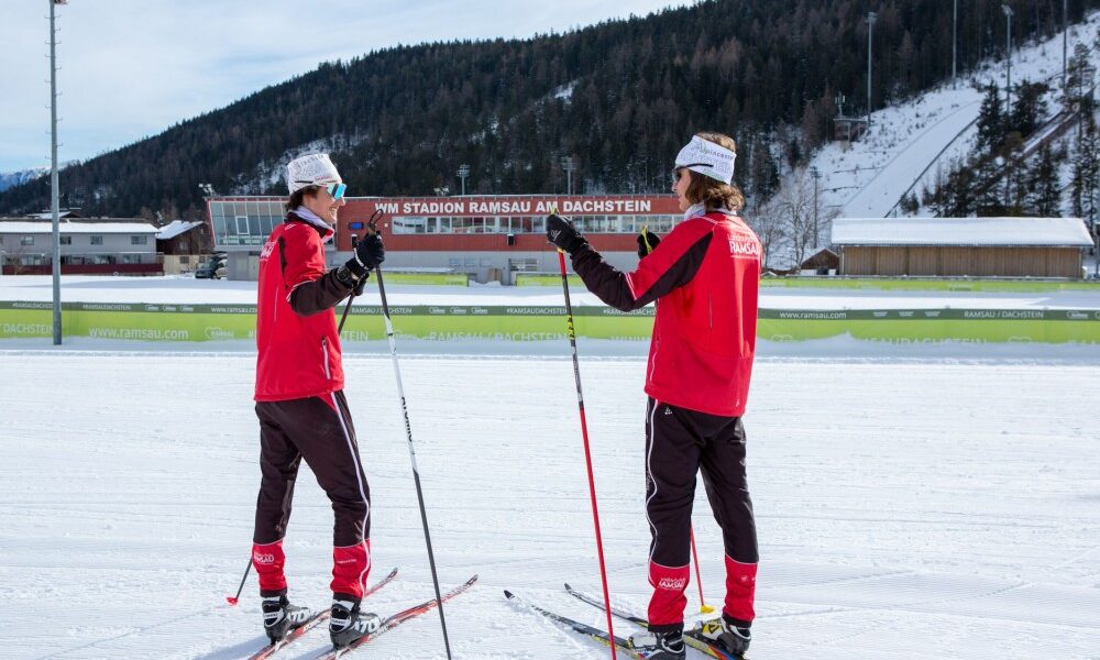 Zwei Skilangläufer in roten Jacken stehen sich auf einer verschneiten Loipe gegenüber, im Hintergrund sind Berge und ein Stadion zu sehen.