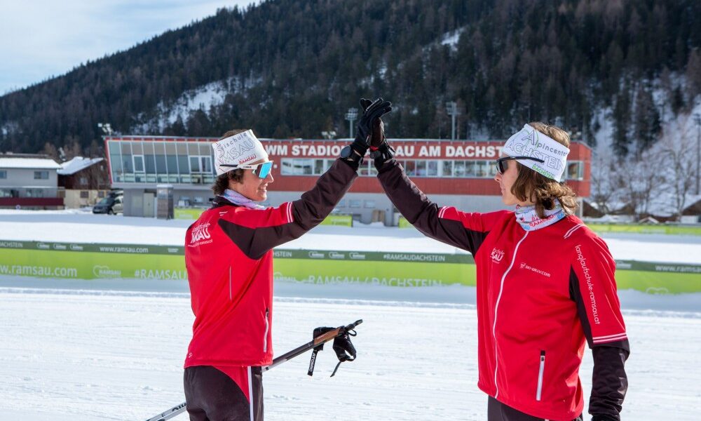Zwei Langläufer in roten Jacken geben sich auf einer verschneiten Strecke vor einem Stadion und schneebedeckten Bergen die Hand.