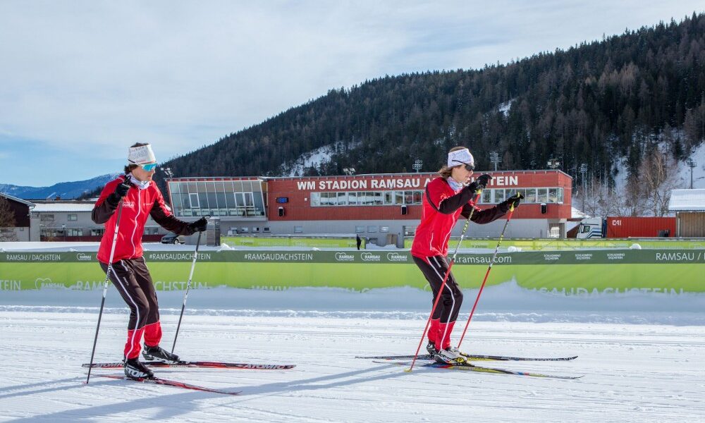 Zwei Langläufer in roten Anzügen fahren auf verschneitem Boden vor dem WM Stadion Ramsau Dachstein, mit Bäumen und Bergen im Hintergrund.