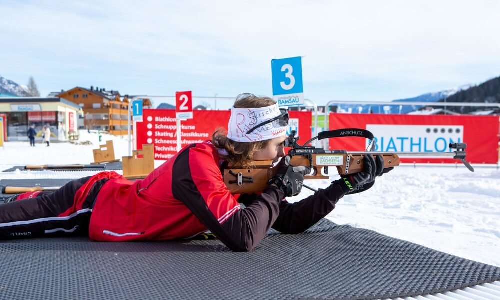 Person in roter Wintersportkleidung, die auf einer Matte liegt und mit einem Gewehr auf einem Biathlon-Schießstand zielt, wobei im Hintergrund Schnee und nummerierte Ziele zu sehen sind.
