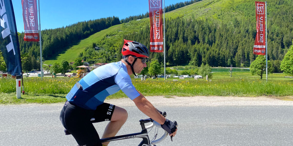 Ein Radfahrer in blauem Trikot und Helm fährt mit seinem Rennrad auf einer asphaltierten Straße, im Hintergrund sind grasbewachsene Hügel, Bäume und Flaggen unter einem klaren blauen Himmel zu sehen.
