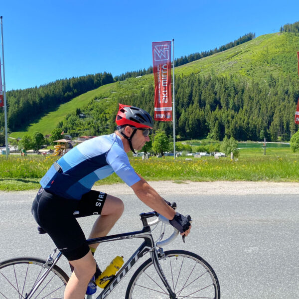 Ein Radfahrer in blauem Trikot und Helm fährt mit seinem Rennrad auf einer asphaltierten Straße, im Hintergrund sind grasbewachsene Hügel, Bäume und Flaggen unter einem klaren blauen Himmel zu sehen.