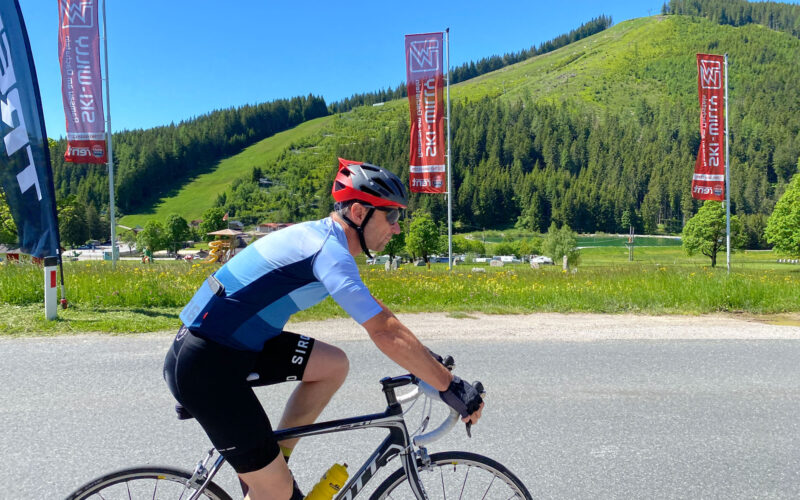 Ein Radfahrer in blauem Trikot und Helm fährt mit seinem Rennrad auf einer asphaltierten Straße, im Hintergrund sind grasbewachsene Hügel, Bäume und Flaggen unter einem klaren blauen Himmel zu sehen.
