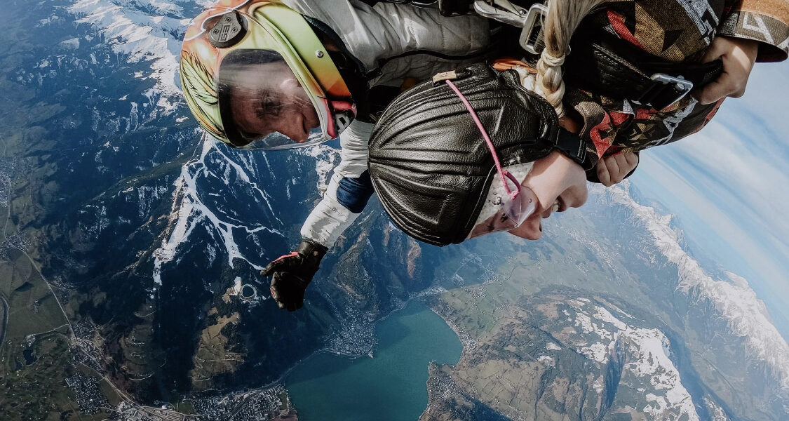 Zwei Personen machen einen Tandem-Fallschirmsprung über einer Berglandschaft, unter der ein See und eine Stadt zu sehen sind; beide tragen Helme und Gurte.