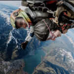 Zwei Personen machen einen Tandem-Fallschirmsprung über einer Berglandschaft, unter der ein See und eine Stadt zu sehen sind; beide tragen Helme und Gurte.