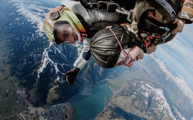 Zwei Personen machen einen Tandem-Fallschirmsprung über einer Berglandschaft, unter der ein See und eine Stadt zu sehen sind; beide tragen Helme und Gurte.