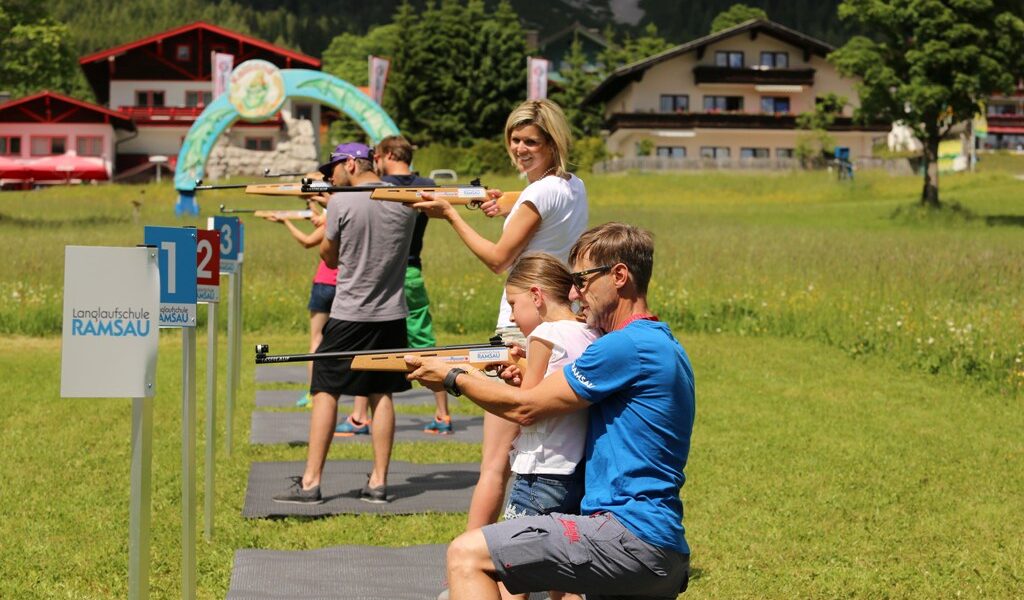 Mehrere Personen, darunter Kinder und Erwachsene, nehmen an einer Outdoor-Aktivität zum Luftgewehrschießen an ausgewiesenen, nummerierten Stationen auf einer Rasenfläche teil.