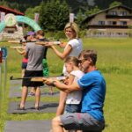Mehrere Personen, darunter Kinder und Erwachsene, nehmen an einer Outdoor-Aktivität zum Luftgewehrschießen an ausgewiesenen, nummerierten Stationen auf einer Rasenfläche teil.