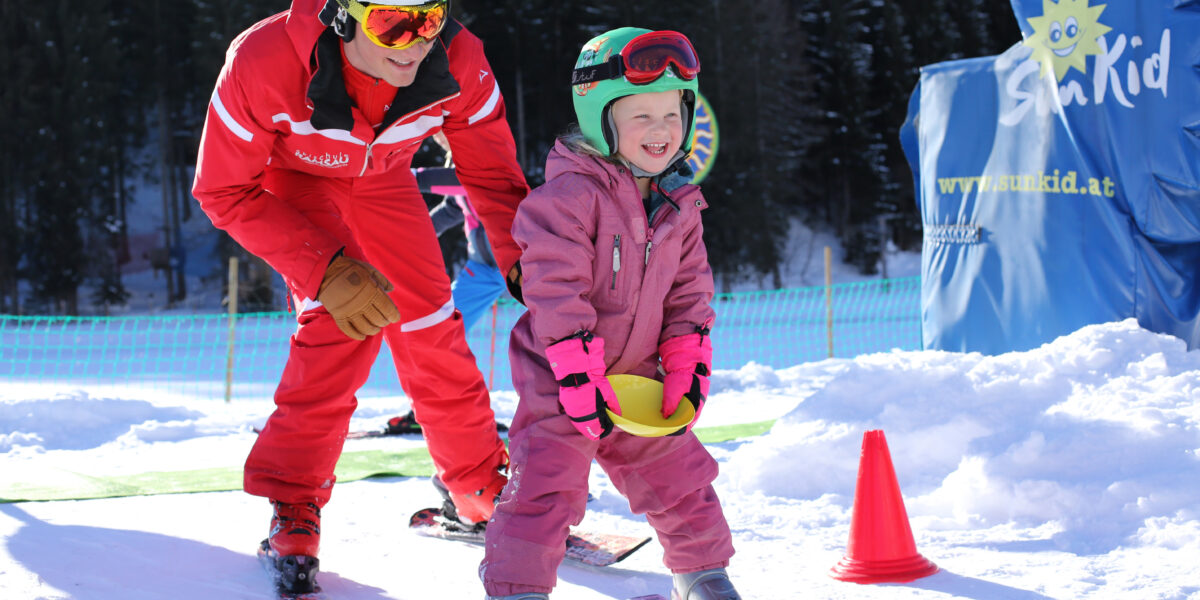 Ein erwachsener Skilehrer hilft einem lächelnden Kind in rosa Skikleidung auf einer verschneiten Piste, mit Sicherheitskegeln und schneebedeckten Bäumen im Hintergrund.