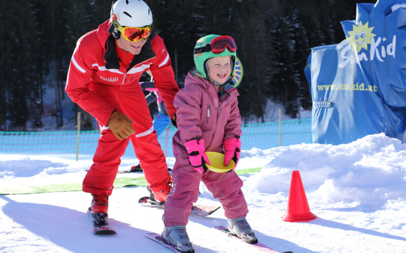 Ein erwachsener Skilehrer hilft einem lächelnden Kind in rosa Skikleidung auf einer verschneiten Piste, mit Sicherheitskegeln und schneebedeckten Bäumen im Hintergrund.