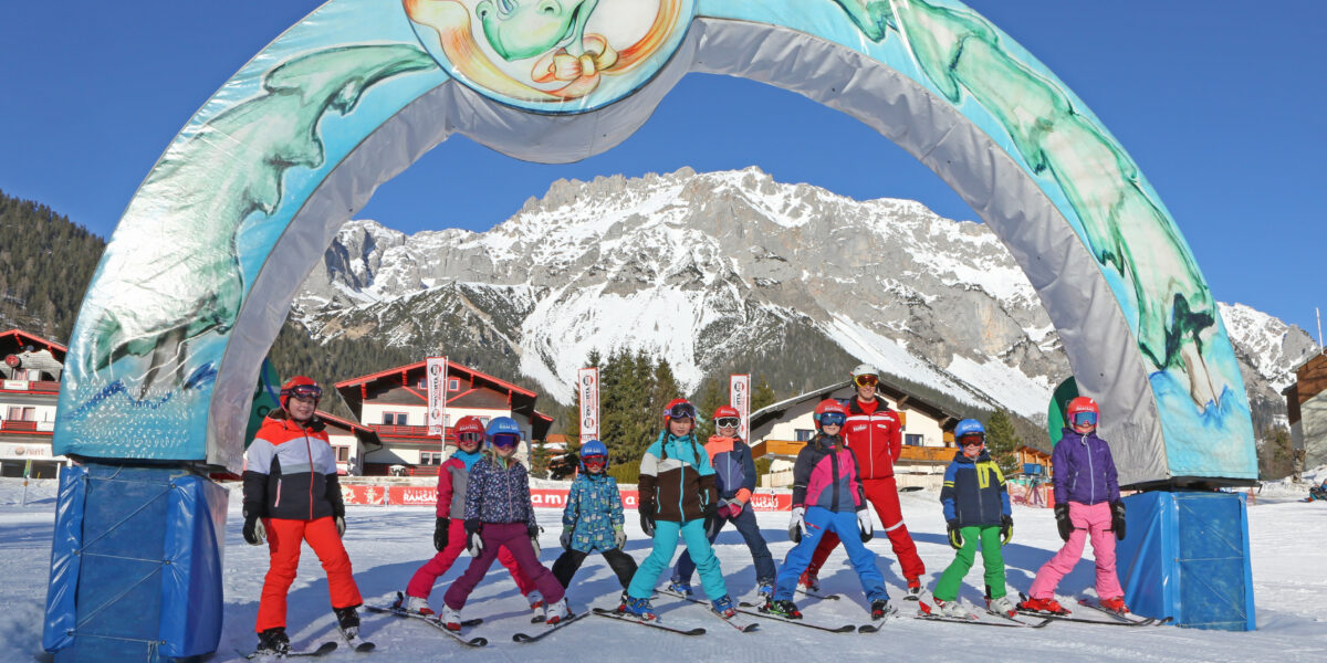 Eine Gruppe von Kindern in bunten Skiklamotten posiert unter einem dekorativen Bogen auf einer verschneiten Skipiste mit Bergen im Hintergrund.