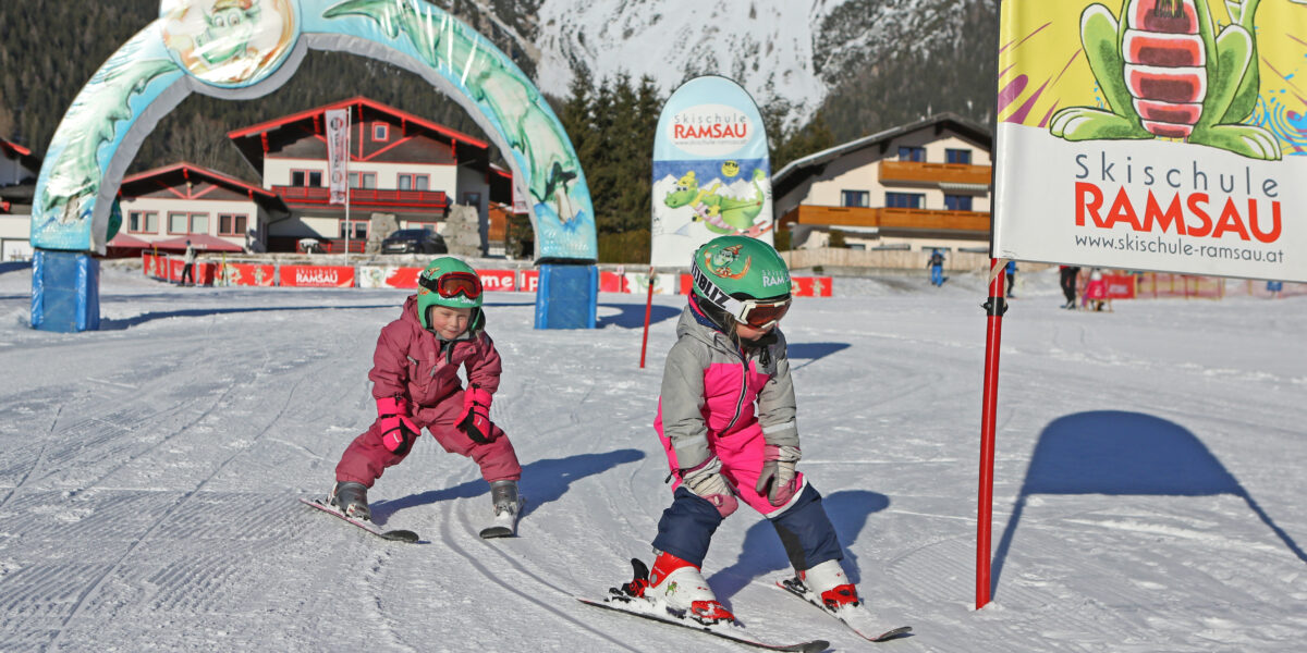 Zwei kleine Kinder in rosafarbenen Schneeanzügen und Helmen fahren auf einer verschneiten Piste neben einem Banner der "Skischule Ramsau" mit Gebäuden und Bergen im Hintergrund Ski.