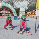 Zwei kleine Kinder in rosafarbenen Schneeanzügen und Helmen fahren auf einer verschneiten Piste neben einem Banner der "Skischule Ramsau" mit Gebäuden und Bergen im Hintergrund Ski.