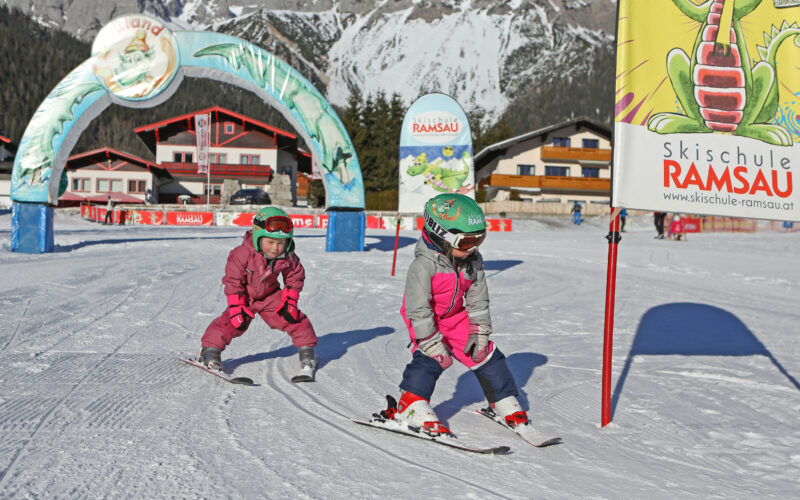 Zwei kleine Kinder in rosafarbenen Schneeanzügen und Helmen fahren auf einer verschneiten Piste neben einem Banner der "Skischule Ramsau" mit Gebäuden und Bergen im Hintergrund Ski.