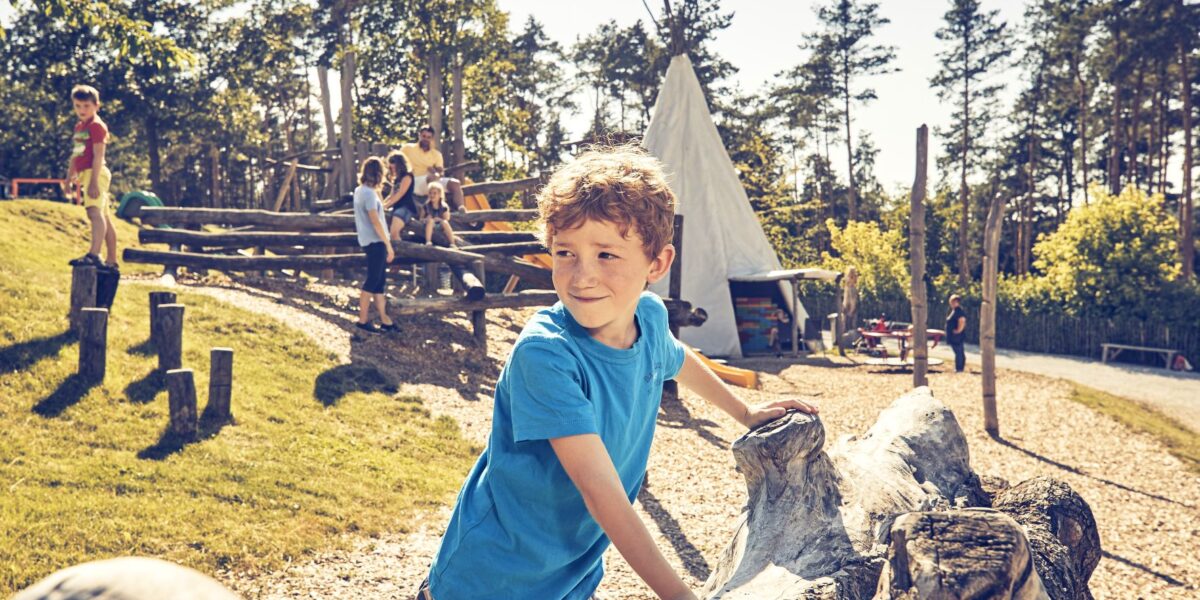 Ein Junge in einem blauen Hemd steht auf Holzstämmen auf einem Spielplatz im Freien; andere Kinder spielen im Hintergrund in der Nähe einer Tipi-Struktur und eines hölzernen Kletterbereichs.