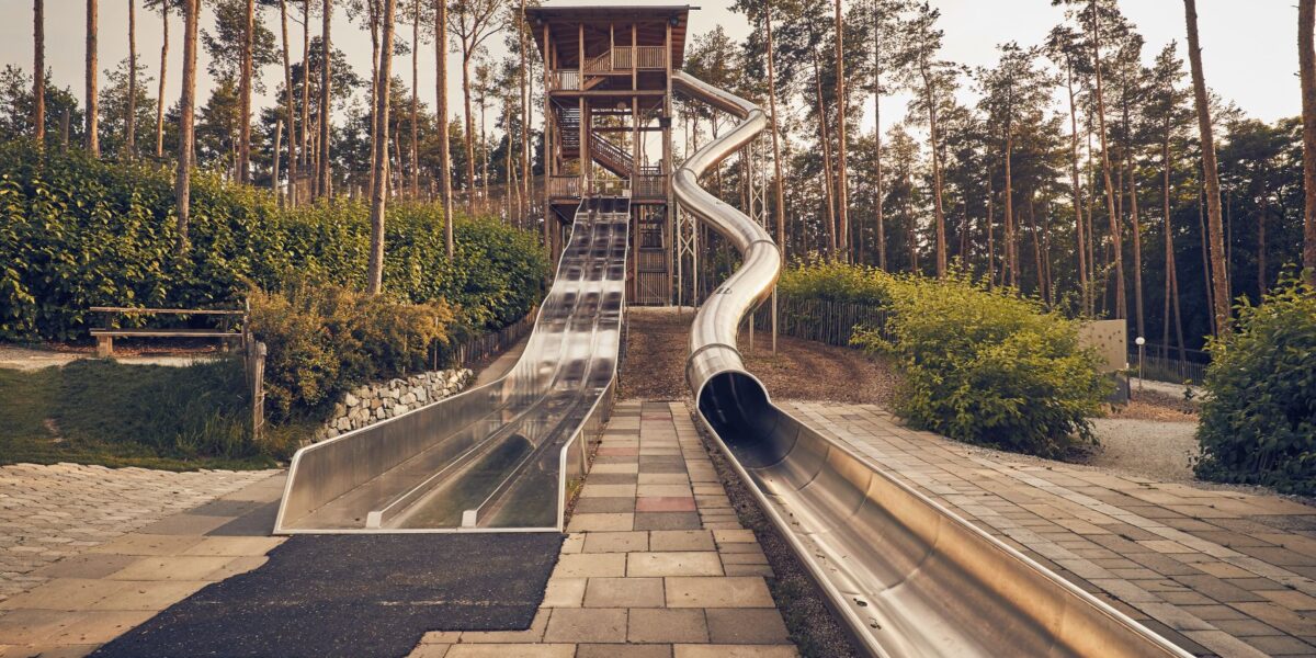 Ein Spielplatz mit drei hohen Metallrutschen nebeneinander, inmitten von Bäumen, mit einer gepflasterten Fläche am Fuß und einer Holztreppe, die nach oben führt.
