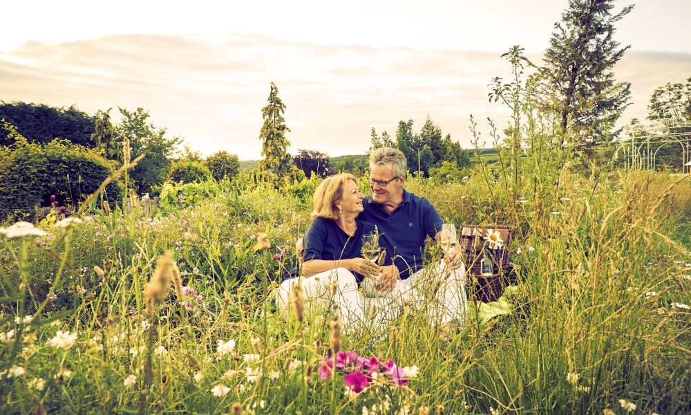 Ein älteres Paar sitzt eng beieinander auf einer Decke auf einer Wiese, umgeben von Wildblumen und hohem Gras, und verbringt einen glücklichen Moment im Freien.