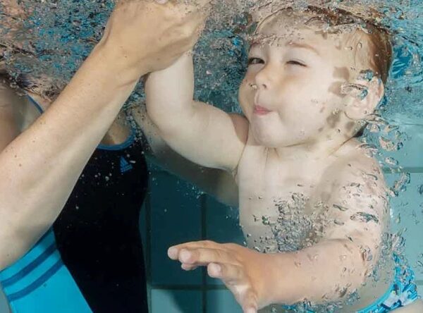 Ein Baby liegt unter Wasser in einem Schwimmbecken und streckt seine Hand nach einem Erwachsenen aus, der es stützt und hält.