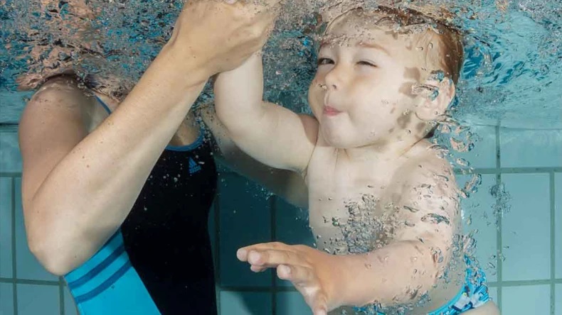 Ein Baby liegt unter Wasser in einem Schwimmbecken und streckt seine Hand nach einem Erwachsenen aus, der es stützt und hält.