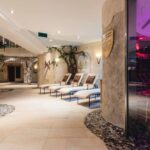 Moderner Indoor-Spa-Bereich mit mehreren Liegestühlen, Steinwänden, Topfpflanzen, gedämpfter Beleuchtung und einer Glastür zu einer Sauna oder einem Dampfbad.