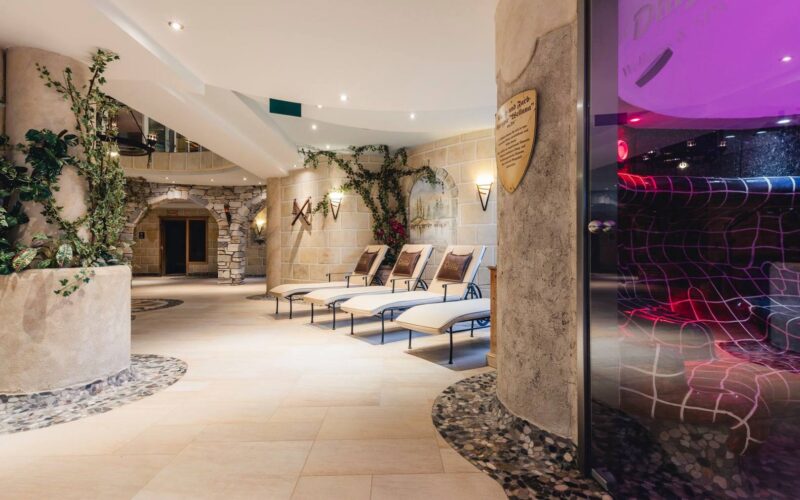 Moderner Indoor-Spa-Bereich mit mehreren Liegestühlen, Steinwänden, Topfpflanzen, gedämpfter Beleuchtung und einer Glastür zu einer Sauna oder einem Dampfbad.