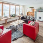 Helles Wohnzimmer mit großen Fenstern, roten Sesseln, dunklen Sofas, einem hölzernen Couchtisch, einem Kamin und einem Blick auf die verschneite Landschaft draußen.