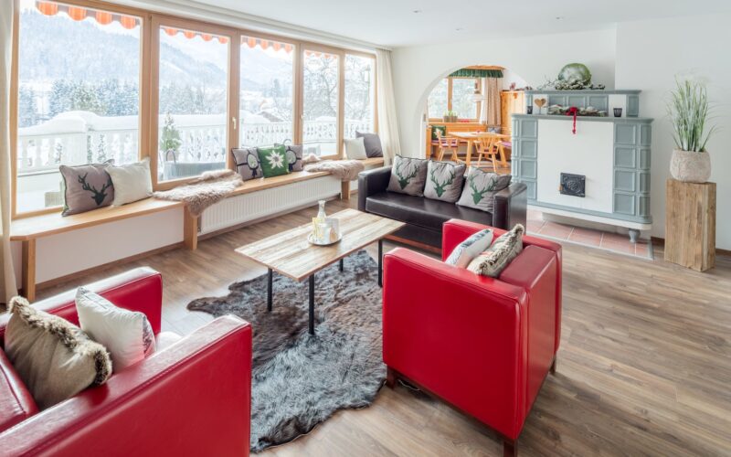 Helles Wohnzimmer mit großen Fenstern, roten Sesseln, dunklen Sofas, einem hölzernen Couchtisch, einem Kamin und einem Blick auf die verschneite Landschaft draußen.