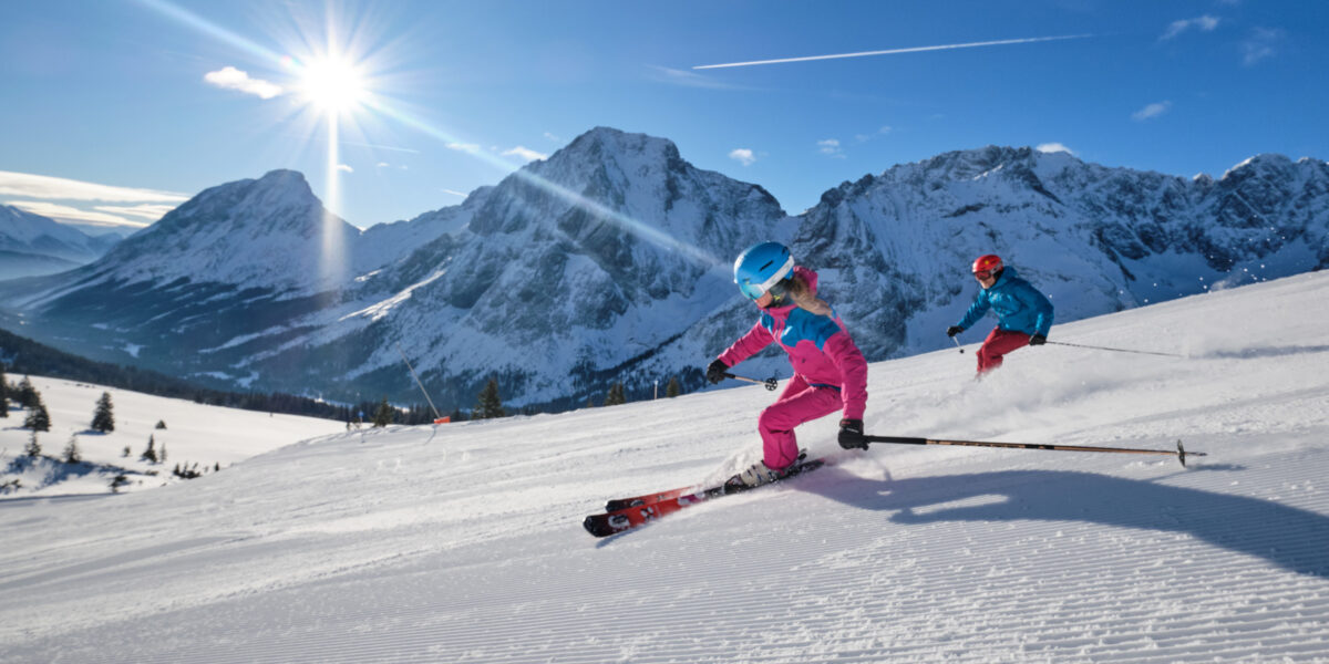 Zwei Personen in farbenfroher Skiausrüstung fahren an einem sonnigen Tag mit schneebedeckten Bergen und einem klaren blauen Himmel im Hintergrund eine präparierte Piste hinunter.