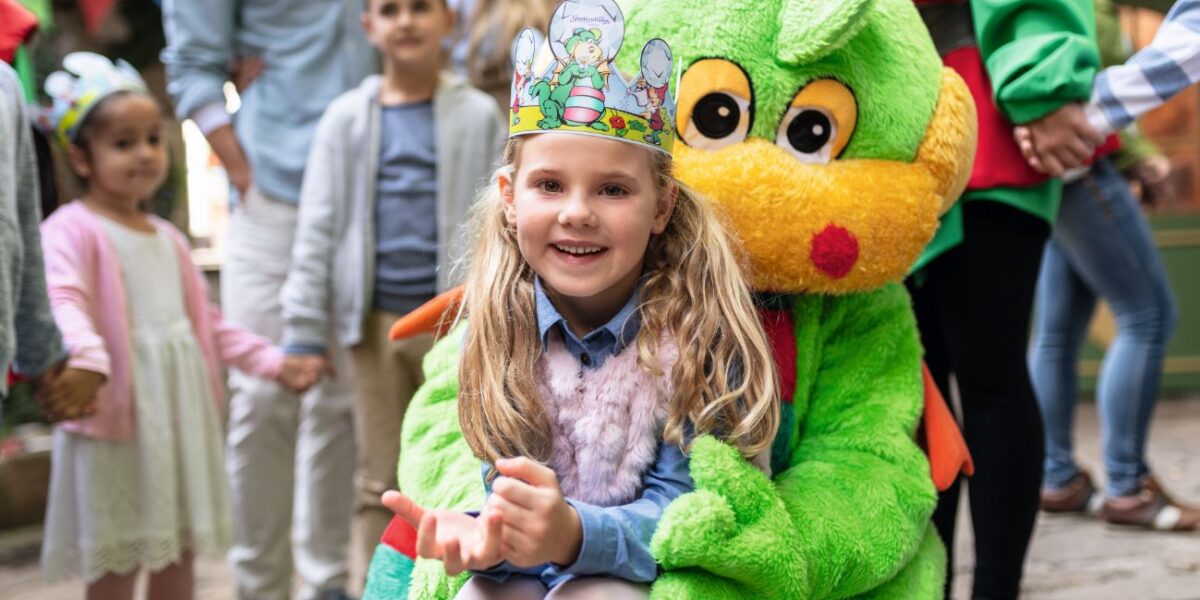 Ein lächelndes Mädchen mit einer Papierkrone sitzt auf dem Schoß einer Person im grünen Drachenkostüm auf einem Kinderfest. Im Hintergrund stehen weitere Kinder und Erwachsene.