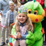 Ein lächelndes Mädchen mit einer Papierkrone sitzt auf dem Schoß einer Person im grünen Drachenkostüm auf einem Kinderfest. Im Hintergrund stehen weitere Kinder und Erwachsene.