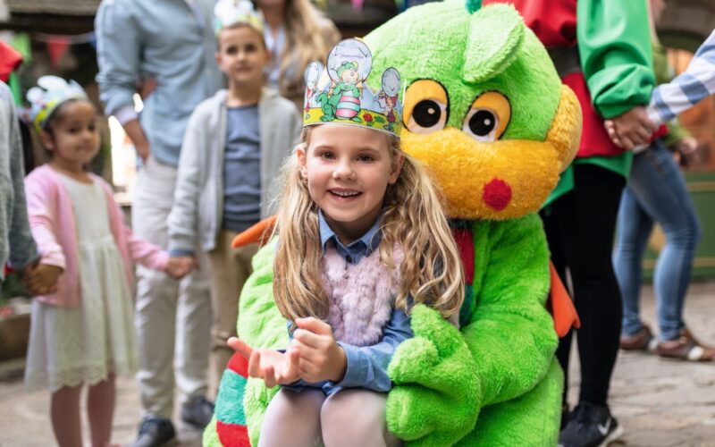 Ein lächelndes Mädchen mit einer Papierkrone sitzt auf dem Schoß einer Person im grünen Drachenkostüm auf einem Kinderfest. Im Hintergrund stehen weitere Kinder und Erwachsene.