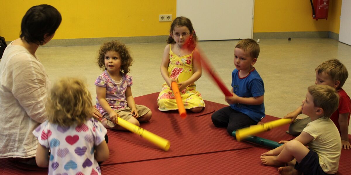 Eine Gruppe kleiner Kinder sitzt im Kreis auf einer Matte und hält bunte Röhren, während ein Erwachsener die Aktivität in einem hellen Raum leitet.