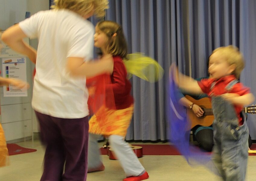 Mehrere Kinder spielen in einem Haus und winken mit bunten Tüchern, während ein Erwachsener im Hintergrund auf einer Gitarre spielt.