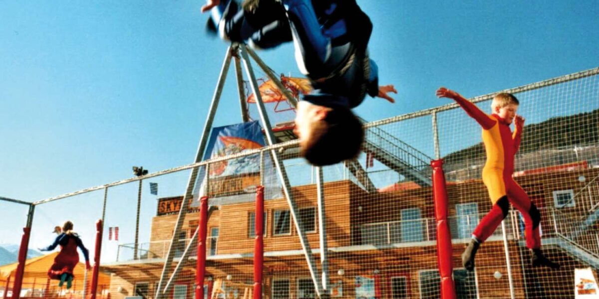 Zwei Kinder in Overalls hüpfen draußen auf einem Trampolin; eines macht gerade einen Salto, während das andere in der Luft ist. Im Hintergrund sind Gebäude und ein Zaun zu sehen.