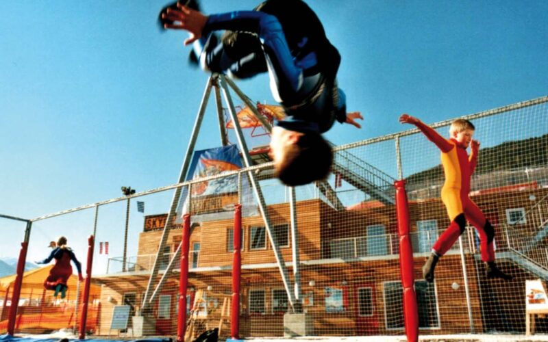 Zwei Kinder in Overalls hüpfen draußen auf einem Trampolin; eines macht gerade einen Salto, während das andere in der Luft ist. Im Hintergrund sind Gebäude und ein Zaun zu sehen.