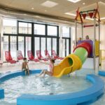 Kinder spielen in einem flachen Hallenbad mit einer gelben Rutsche und Wasserspielen; rote Liegestühle stehen im Hintergrund neben großen Fenstern.