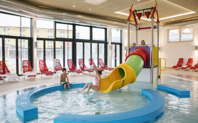 Kinder spielen in einem flachen Hallenbad mit einer gelben Rutsche und Wasserspielen; rote Liegestühle stehen im Hintergrund neben großen Fenstern.