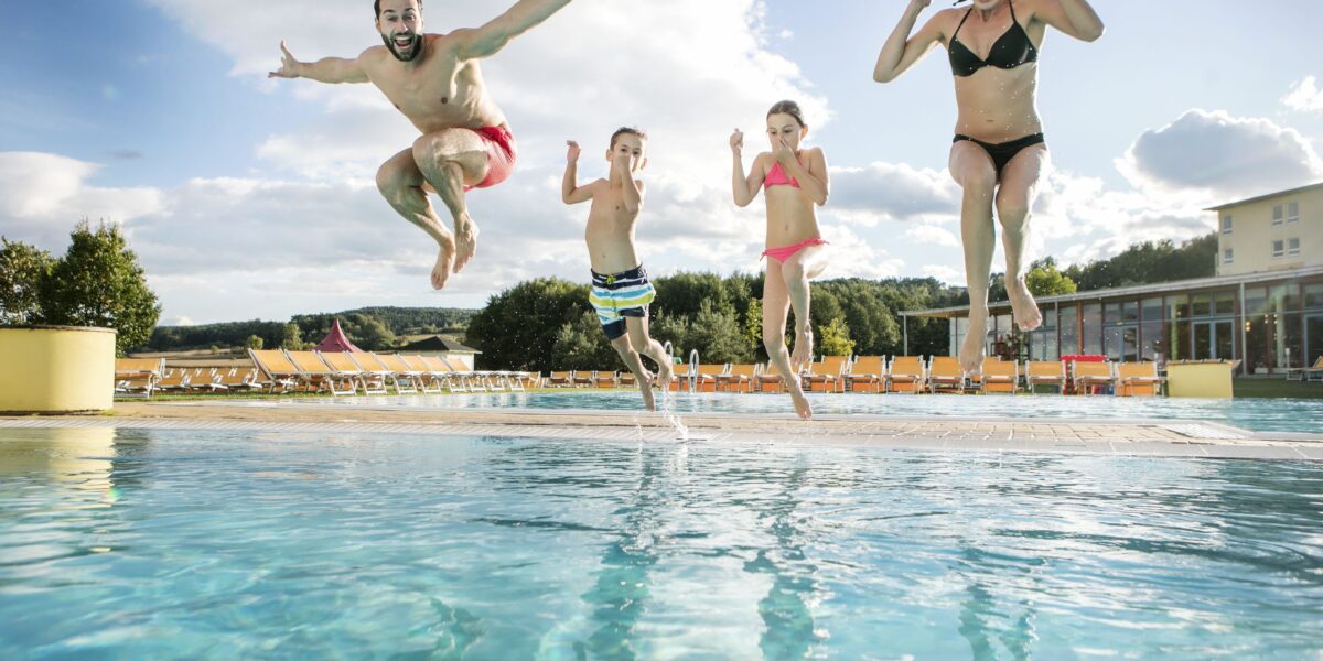 Vier Personen, zwei Erwachsene und zwei Kinder, springen an einem sonnigen Tag gemeinsam in ein Freibad.