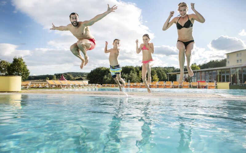 Vier Personen, zwei Erwachsene und zwei Kinder, springen an einem sonnigen Tag gemeinsam in ein Freibad.