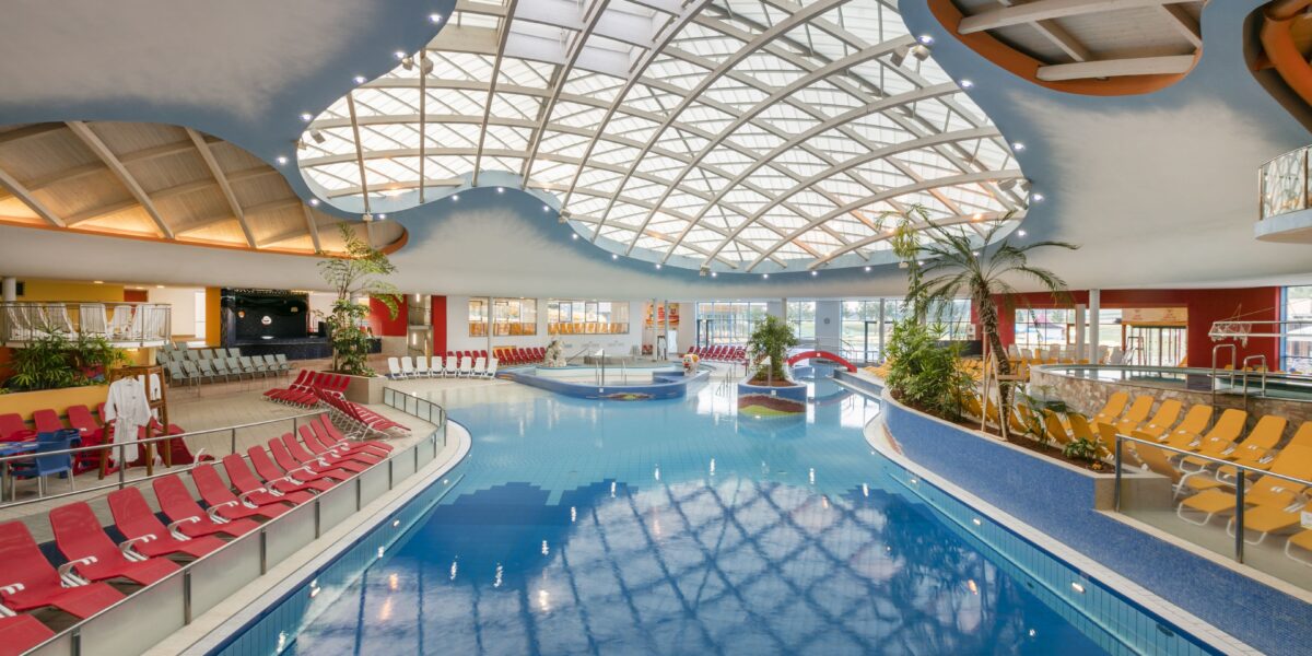 Hallenbad mit roten Liegestühlen, Palmen und einer großen geschwungenen Glasdecke, die natürliches Licht in den großzügigen Bereich lässt.