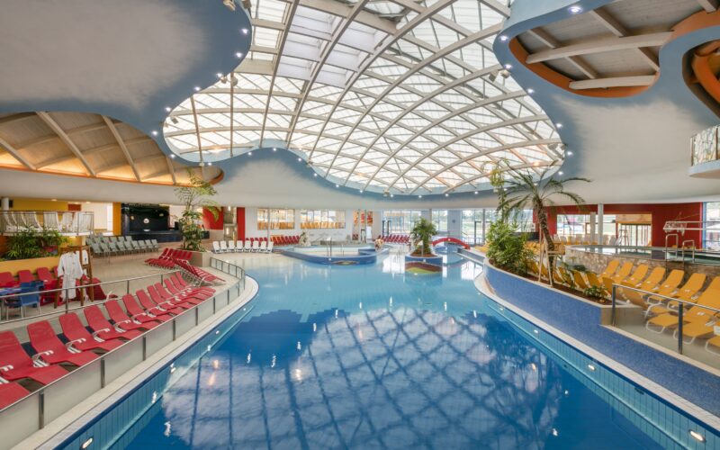 Hallenbad mit roten Liegestühlen, Palmen und einer großen geschwungenen Glasdecke, die natürliches Licht in den großzügigen Bereich lässt.