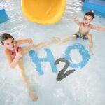 Zwei Kinder in Badeanzügen sitzen in einem flachen Becken neben einer gelben Rutsche und zeigen mit dem auf den Beckenboden gemalten "H2O" die Daumen nach oben.