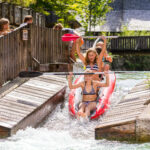 Drei Mädchen in Badeanzügen fahren in einem roten Kajak eine schmale Wasserrutsche hinunter und planschen, während sie in einer sonnigen Umgebung im Freien an Holzgeländern und Zuschauern vorbeikommen.