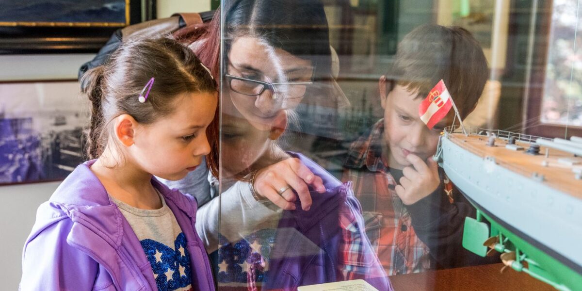 Eine Frau und zwei Kinder sehen sich eine Museumsausstellung mit einem Schiffsmodell hinter Glas genau an und lesen eine Informationstafel.