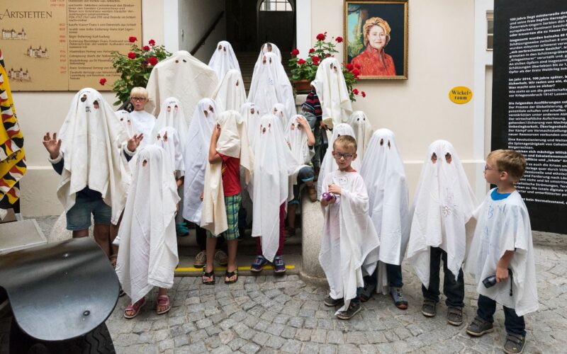 Eine Gruppe von Kindern und Erwachsenen, die als Gespenster mit weißen Tüchern verkleidet sind, posieren gemeinsam in einem Innenhof.